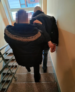 Zdjęcie przedstawia policjanta ubranego w czarną kurtkę z kapturem obszytym brązowym futrem, który sprowadza po schodach zatrzymanego mężczyznę. Mężczyzna ten jest ubrany w czarną kurtkę, na rękach trzymanych z tyłu ma założone kajdanki. Obaj znajdują się tyłem do obiektywu aparatu.