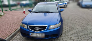 Zdjęcie przedstawia niebieski pojazd m-ki Honda, który stoi prosto do obiektywu aparatu. Jego tablice rejestracyjne są zasłonięte. Za nim widać zaparkowany oznakowany radiowóz.