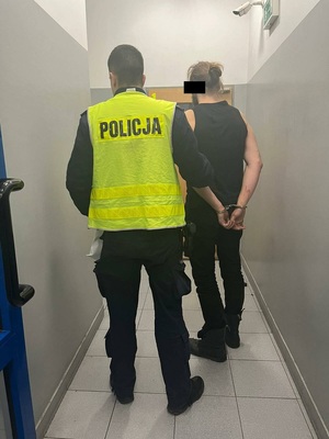 Zdjęcie przedstawia policjanta ubranego w odblaskową kamizelkę z napisem POLICJA, który prowadzi przez korytarz zatrzymanego policjanta. Obaj są tyłem do obiektywu aparatu. Zatrzymany mężczyzna stoi po prawej stronie policjanta. Mężczyzna ma dłuższe włosy spięte w kucyk, jest ubrany na ciemno. Na rękach trzymanych z tyłu ma założone kajdanki.