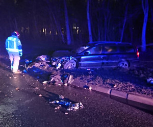 Zdjęcie przedstawia miejsce kolizji, jest ciemno. widać na zdjęciu rozbity szary samochód, po lewej stronie zdjęcia widać strażaka.