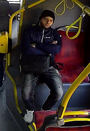 Zdjęcie przedstawia siedzącego w autobusie, przodem do kamery mężczyznę. Ma około 25-30 lat, jest wysokiego wzrostu, ma wysportowaną sylwetkę, posiada ciemny zarost. Ubrany jest w czarną kurtkę, ciemne spodnie jeansowe, czarną wełnianą czapkę i białe buty.