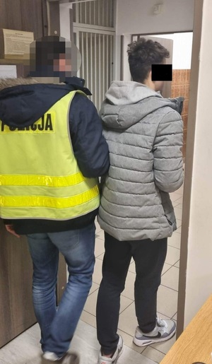 Zdjęcie przedstawia policjanta w odblaskowej kamizelce z napisem POLICJA, który prowadzi po swojej prawej stronie zatrzymanego nastolatka. Część twarzy chłopaka zasłania czarny prostokąt. Jest on ubrany w szarą kurtkę zimową.