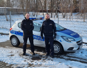 Zdjęcie przedstawi dwóch umundurowanych policjantów, którzy stoją przed zaparkowanym bokiem radiowozem.