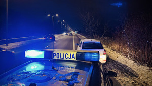 Zdjęcie przedstawia oznakowany radiowóz z włączonymi światłami błyskowymi, który stoi za zaparkowanym białym pojazdem na poboczu drogi.