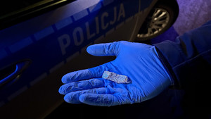 Zdjęcie przedstawia dłoń w niebieskiej rękawiczce ochronnej, na której leży małe zawiniątko. w Tle widać oznakowany radiowóz policyjny.