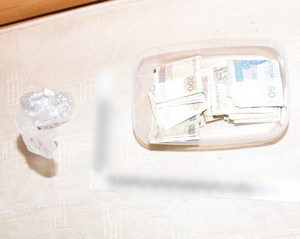 Zdjęcie przedstawia przezroczystą torebkę foliową zawierającą srebrne zawiniątka, która leży na materacu. Po prawej stronie widać plastikowy przezroczysty pojemnik z banknotami w różnych nominałach.