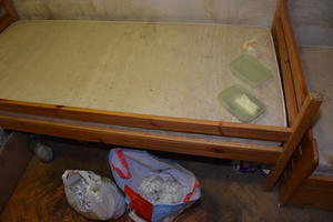 Zdjęcie przedstawia pomieszczenie, w którym stoi łóżko. Na nim widać plastikowy przezroczysty pojemnik, a przed łóżkiem stoją dwie torby w których znajdują się srebrne zawiniątka.