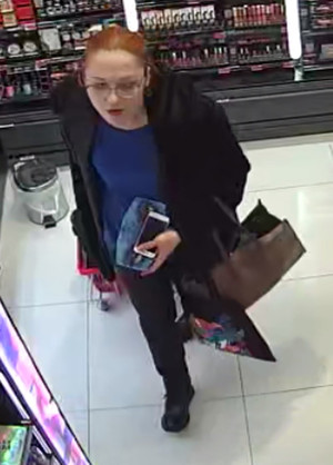 Zdjęcie przedstawia kobietę w ciemnej kurtce, niebieskim swetrze, okularach korekcyjnych, która znajduje się wewnątrz drogerii. Wokół niej widać półki z asortymentem. W lewej dłoni trzyma jakiś kwadratowy przedmiot. Przez lewą rękę ma przełożoną brązową torebkę.
