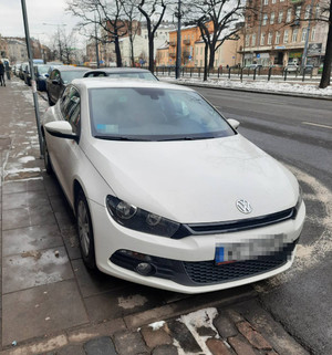Zdjęcie przedstawia biały pojazd marki Volkswagen zaparkowany przy ulicy. Ma on zasłonięte tablice rejestracyjne i nalepkę legalizacyjną. W tle widać inne zaparkowane przy ulicy pojazdy.