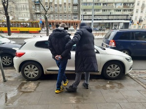 Zdjęcie przedstawia zaparkowany przy ulicy samochód osobowy w jasnym kolorze. Stoi przy nim osoba ubrana w ciemny długi płaszcz i przytrzymuje za rękę stojącego po jego lewej stronie mężczyznę, który ma kajdanki założone na ręce trzymane z tyłu. Mężczyzna ten jest ubrany na ciemno, ma buty sportowe w kolorowych odcieniach. W tle widać tramwaj i sklepy.