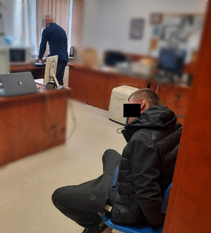 Zdjęcie przedstawia ubranego na ciemno mężczyznę, który siedzi na krześle w pomieszczeniu tuż przy szafie. Jego twarz zasłania czarny prostokąt. Po drugiej stronie widać ubranego na ciemno policjanta. Jego twarz jest zasłonięta komputerowym filtrem.