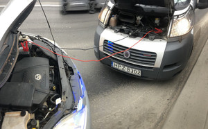 Zdjęcie przedstawia oznakowany radiowóz policji oraz samochód osobowy stojące naprzeciwko siebie na pasie ruchu. Obaj auta mają podniesione maski, spod których widać kolorowe kable przeciągnięte od jednego do drugiego.