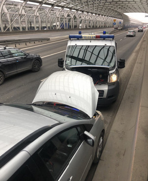Zdjęcie przedstawia oznakowany radiowóz policji oraz samochód osobowy stojące naprzeciwko siebie na pasie ruchu. Obaj auta mają podniesione maski, spod których widać kolorowe kable przeciągnięte od jednego do drugiego.