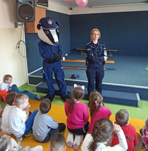 Zdjęcie przedstawia umundurowaną policjantkę i osobę przebraną w strój maskotki Borsuka, którzy stoją przed dziećmi siedzącymi na dywanie.
