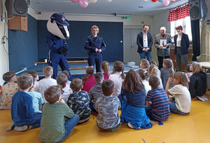 Zdjęcie przedstawia umundurowaną policjantkę, osobę przebraną w strój maskotki Borsuka oraz trzy inne osoby, które stoją przed dziećmi siedzącymi na dywanie.