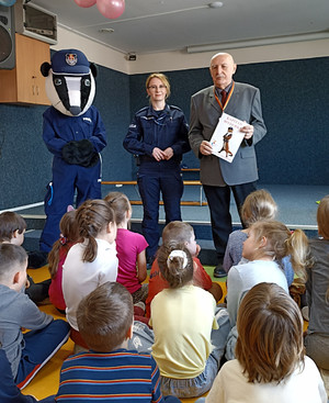 Zdjęcie przedstawia umundurowaną policjantkę, osobę przebraną w strój maskotki Borsuka oraz mężczyznę, którzy stoją przed dziećmi siedzącymi na dywanie.