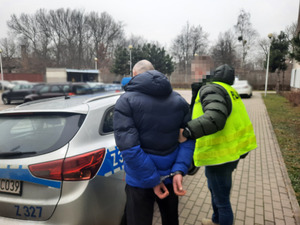 Zdjęcie przedstawia policjanta ubranego w odblaskową kamizelkę z czarnym napisem POLICJA na plecach, który stoi z zatrzymanym mężczyzną przy radiowozie. Mężczyzna jest ubrany w granatowo-czarną kurtkę puchową i ciemne spodnie. Na rękach trzymanych z tyłu ma założone kajdanki. Twarz policjanta jest zasłonięta. Obaj stoją lewym profilem do obiektywu aparatu.