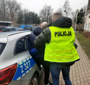 Zdjęcie przedstawia policjanta ubranego w odblaskową kamizelkę z czarnym napisem POLICJA na plecach, który pomaga wsiąść do radiowozu zatrzymanemu mężczyźnie. Mężczyzna jest ubrany w granatowo-czarną kurtkę puchową i ciemne spodnie. Na rękach trzymanych z tyłu ma założone kajdanki. Twarz policjanta jest zasłonięta.