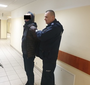Zdjęcie przedstawia umundurowanego policjanta, kory stoi na korytarzu razem z zatrzymanym mężczyzną. Mężczyzna stoi po prawej stronie policjanta. Trzyma ręce z tyłu. Na głowie ma założoną kominiarkę. Część jego twarzy zasłania czarny prostokąt. Obaj stoją lewym profilem do obiektywu aparatu.
