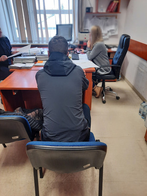 Zdjęcie przedstawia policjantkę ubraną w cywilne ubrania, która siedzi przed monitorem komputera, lewym profilem do obiektywu aparatu. Zatrzymany mężczyzna siedzi przy innym biurku, tyłem do wejścia do pokoju. Mężczyzna jest ubrany na ciemno.