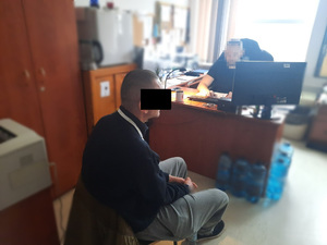 Zdjęcie przedstawia policjanta ubranego na ciemno, który siedzi za biurkiem przed monitorem komputera, przodem do wejścia do pokoju i obiektywu aparatu.
Prawym profilem do zdjęcia, naprzeciwko biurka za którym siedzi policjant siedzi zatrzymany mężczyzna. Jego twarz jest zasłonięta czarnym prostokątem. Mężczyzna jest ubrany w czarną bluzę i szare spodnie. Dłonie trzyma razem na kolanach.