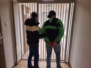 Zdjęcie przedstawia policjanta ubranego w czarną kurtkę z białym paskiem na wysokości klatki piersiowej i ciemne spodnie, który stoi przed okratowanymi drzwiami pomieszczenia razem z zatrzymanym mężczyzną. Twarz policjanta jest rozmazana komputerowym filtrem. Po jego prawej stronie stoi zatrzymany mężczyzna. Jest on ubrany w zielono-czarną kurtkę puchową, czarną czapkę z daszkiem i szare spodnie. Na rekach trzymanych z tyłu ma założone kajdanki. Część jego twarzy zasłania czarny prostokąt. Obaj stoją tyłem do obiektywu aparatu.