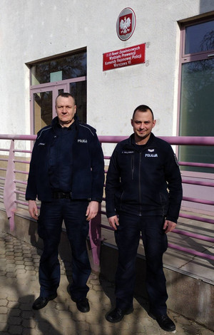 Zdjęcie przedstawia dwóch umundurowanych policjantów, którzy stoją przodem do obiektywu aparatu. Stoją oni na tle drzwi prowadzących do budynku, nad którym znajduje się czerwoną tabliczka z białym napisem.