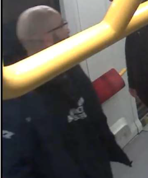 Zdjęcie przedstawia fragment sylwetki mężczyzny o bardzo krótkich włosach. Ma on czarną bluzę z białym napisem na wysokości klatki piersiowej. Nad jego głową widać żółtą poręcz do przytrzymywania się podczas jazdy metrem.