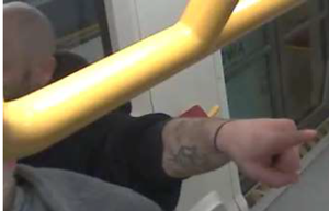 Zdjęcie przedstawia fragment sylwetki mężczyzny o bardzo krótkich włosach. Ma on czarną bluzę. Na lewym przedramieniu ma tatuaż.