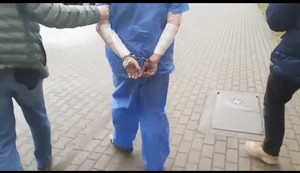 Zdjęcie przedstawia fragmenty sylwetek policjantów, który prowadzą ubranego w odzież ochronną niebieską zatrzymanego mężczyznę. Ma on bandaże na przedramionach. Ręce trzyma z tyłu w kajdankach.