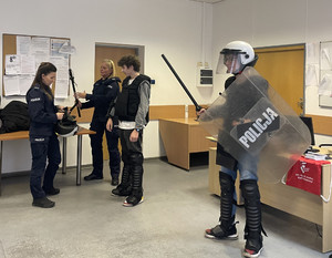 Na zdjęciu widać dwie umundurowane policjantki oraz dwie osoby wyposażone w sprzęt policyjny wykorzystywane podczas zabezpieczeń- tj. kask, pałka, tarcza, kamizelkę ochraniającą klatkę piersiową