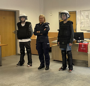 Na zdjęciu widać umundurowaną policjantkę po bokach której stoją dwie osoby ubrane w sprzęt policyjny wykorzystywany podczas zabezpieczeń