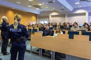 Zdjęcie przedstawia dwie umundurowane policjantki, które stoją tyłem do obiektywu aparatu przed siedzącymi w ławkach studentami.