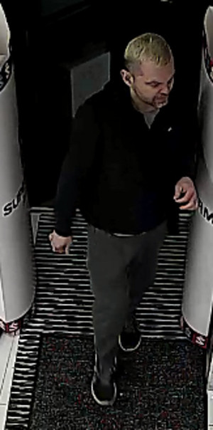Zdjęcie przedstawia mężczyznę wchodzącego do środka drogerii przez bramki alarmowe. Jest on w wieku około 30-35 lat, ma około 175 cm wzrostu, przeciętną budowę ciała, włosy koloru blond; ubrany jest w czarną bluzę polarową, ciemne spodnie i ciemne sportowe buty. Jest skierowany przodem do kamery monitoringu.