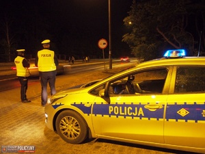 Zdjęcie przedstawia oznakowany radiowóz zaparkowany przy drodze, przy którym stoi dwóch umundurowanych policjantów. Zdjęcie jest wykonane, gdy na zewnątrz jest ciemno.