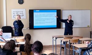 Zdjęcie przedstawia dwie umundurowane policjantki, które stoją po obu stronach prezentacji wyświetlanej na planszy na ścianie przed siedzącymi w ławkach uczniami.