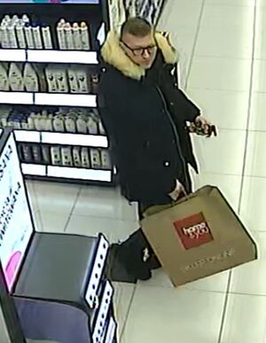 Mężczyzna jest w wieku około 35-40 lat, wysokiego wzrostu, ma przeciętną budowę ciała, włosy krótkie ciemne, okulary korekcyjne; ubrany jest w długą, zimową i ciemną kurtkę z futrzanym obszyciem, ciemnoniebieskie spodnie jeansowe, czarne buty zimowe; posiada papierową torbę z napisem "Home&amp;You". W tle widać półki z asortymentem sklepowym.