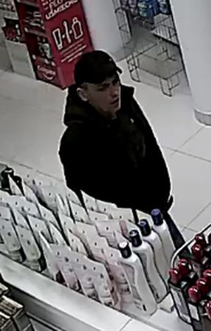 Zdjęcie przedstawia mężczyznę idącego po sklepie. Ma on około 30 lat; ubrany jest w czarną czapkę z daszkiem, czarną kurtkę. Wokół niego widać półki z asortymentem sklepowym.