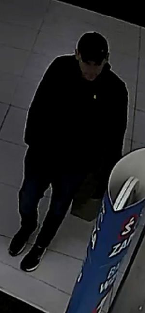 Zdjęcie przedstawia mężczyznę idącego po sklepie. Ma on około 30 lat, około 180 cm wzrostu, szczupłą budowę ciała, ubrany jest w czarną czapkę z daszkiem, czarną kurtkę, spodnie w ciemnym kolorze, sportowe buty z białą podeszwą, w lewej dłoni trzyma papierową torbę zakupową z białym napisem „Reserved”. Wokół niego widać półki z asortymentem sklepowym.