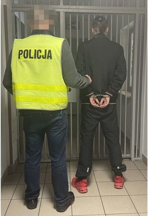 Zdjęcie przedstawia nieumundurowanego policjanta ubranego w odblaskową kamizelkę, który wyprowadza z pomieszczenia dla zatrzymanych mężczyznę, który ma na rękach trzymanych z tyłu kajdanki.