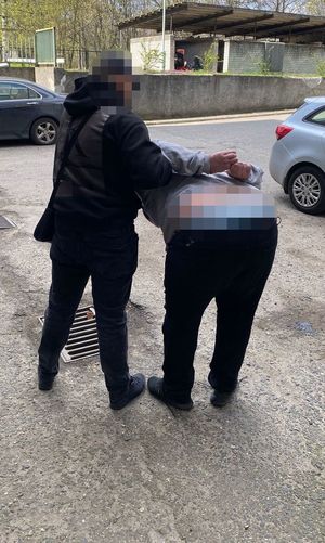 Zdjęcie prowadzi policjanta w cywilnych ubraniach, który prowadzi zatrzymanego mężczyznę. Mężczyzna ten jest pochylony do przodu. Na rekach trzymanych z tyłu ma założone kajdanki.