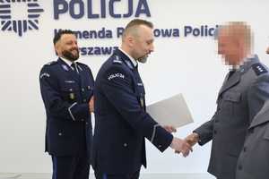 Zdjęcie przedstawia policjantów w galowych mundurach, którzy stoją w jednym rzędzie. Przed policjantem stoi jej przełożony, który podaje jej rękę. W tle widać białą ścianę z granatowym napisem KOMENDA REJONOWA POLICJI WARSZAWA VI.