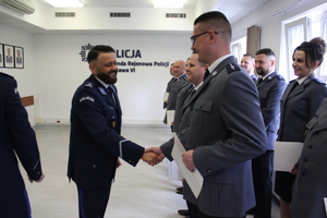 Zdjęcie przedstawia policjantów w galowych mundurach, którzy stoją w jednym rzędzie. Przed policjantem sto jego przełożony, który podaje mu rękę. W tle widać białą ścianę z granatowym napisem KOMENDA REJONOWA POLICJI WARSZAWA VI.