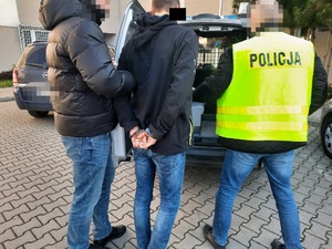 Zdjęcie przedstawia osobę ubraną na ciemno, która prowadzi po swojej prawej stronie zatrzymanego mężczyznę. Mężczyzna ten ma kajdanki założone na rękach trzymanych z tyłu. Po jego prawej stronie widać osobę ubraną w cywilną odzież i odblaskową kamizelkę z napisem na plecach POLICJA. Wszyscy stoją tyłem do obiektywu aparatu. Przed nimi widać otwarte drzwi tylne oznakowanego radiowozu. W tle widać budynek komendy z okratowanymi oknami.