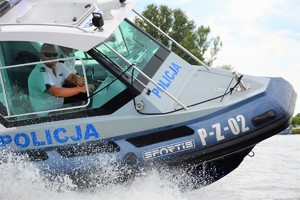 Zdjęcie przedstawia osobę w białej koszulce, która steruje policyjną motorówką.