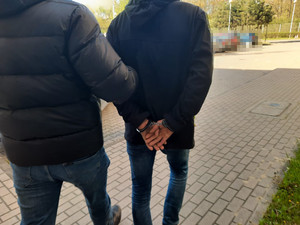 Zdjęcie przedstawia dwie sylwetki mężczyzn. Ten idący po prawej stronie ma kajdanki założone na rekach trzymanych z tyłu.