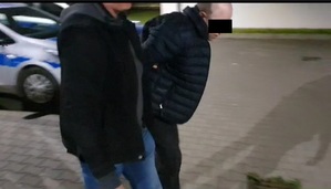 Zdjęcie przedstawia ubranego na ciemno policjanta, który prowadzi po swojej lewej stronie zatrzymanego mężczyznę. Mężczyzna ten jest pochylony do przodu. Część twarzy, na wysokości oczu zasłania mu czarny prostokąt. W tle, za ich plecami widać zaparkowany oznakowany radiowóz. Zdjęcie jest wykonane nocą.