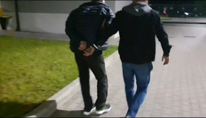 Zdjęcie przedstawia ubranego na ciemno policjanta, który prowadzi po swojej lewej stronie zatrzymanego mężczyznę. Mężczyzna ten ma kajdanki założone na rekach trzymanych z tyłu. Przed nimi widać budynek.