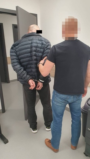 Zdjęcie jest zrobione w pomieszczeniu. Przedstawia ubranego w ciemną koszulkę na krótki rękaw i niebieskie spodnie jeansowe policjanta, który prowadzi przed sobą zatrzymanego mężczyznę. Mężczyzna ten ma kajdanki założone na rekach trzymanych z tyłu. Jest ubrany w czarną pikowaną kurtkę i ciemne spodnie. Część twarzy zasłania mu czarny prostokąt. Obaj na zdjęciu stoją tyłem.