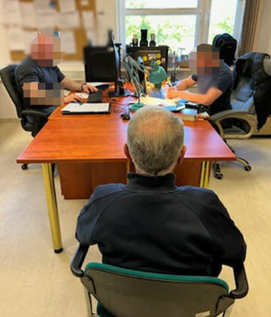 Zdjęcie przedstawia mężczyznę siedzącego na krześle tyłem do wejścia. Przed nim przy biurku siedzi dwóch policjantów, ich twarze są zasłonięte.